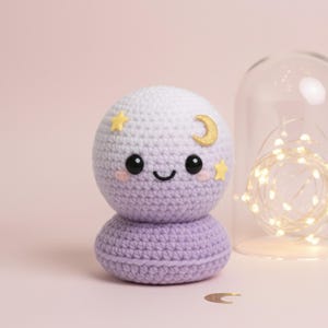May include: A handcrafted crochet moon doll with a smiling face and star details. The doll is in shades of lavender and white, with a gold crescent moon and yellow star accents. A ball of lavender yarn and a glass dome with fairy lights are in the background.