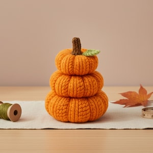 May include: A stack of three orange knitted pumpkins with a brown stem and a green leaf. A spool of green thread, a maple leaf, and a measuring tape are also present, suggesting a fall or harvest theme.