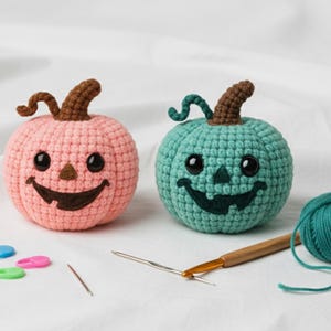 Peut inclure: Deux citrouilles crochetées, une rose et une turquoise, avec des visages souriants de jack-o'-lantern et des tiges marron. Une pelote de fil turquoise, un crochet et des aiguilles sont également présents, suggérant un projet artisanal. Les citrouilles sont faites à la main.