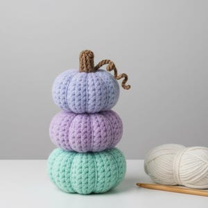 May include: A stack of three crocheted pumpkins in pastel colors: light blue, purple, and teal. A brown stem and curly vine top the pumpkins. Two balls of white yarn and a wooden crochet hook are next to the pumpkins.
