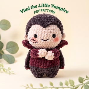 May include: A crocheted vampire doll with black hair, a pale face, black eyes, and a single white fang. The doll wears a dark burgundy cape and matching shirt, with dark grey pants. The text 'Vlad the Little Vampire PDF PATTERN' is visible.