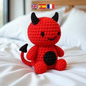 Puede incluir: Muñeca de diablo roja de ganchillo hecha a mano con cuernos negros, una cola negra y una mancha negra en la barriga. La muñeca tiene una cara sonriente con ojos negros. La muñeca está sentada en una cama blanca.