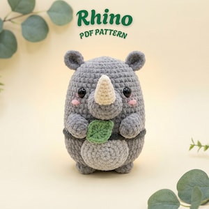 May include: A gray crocheted rhino plush toy with a cream-colored horn, black safety eyes, and pink cheeks, holding a small green leaf. The text Rhino PDF PATTERN is displayed above the toy in dark green letters against a light background.