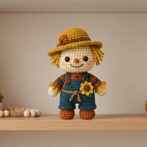 Op de afbeelding: Een handgemaakte gehaakte vogelverschrikkerpop met een gele hoed, strohaar en een zonnebloem op de tuinbroek. De pop draagt een blauwe tuinbroek met oranje en bruine details. De pop is ongeveer 18 cm hoog.