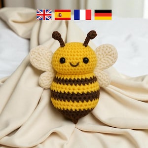 May include: A crocheted bee toy in yellow and brown, with cream wings and small antennae. The bee has a smiling face with black eyes. The toy is set against a cream-coloured fabric background. The bee is approximately 10 cm in height.