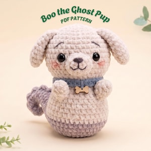 Op de afbeelding: Een gehaakt knuffeltje in de vorm van een puppy van lichtbeige garen met hangende oren, zwarte veiligheidsoogjes en roze wangen. Het draagt een blauwe halsband met een klein houten botvormig label. De tekst erboven luidt Boo the Ghost Pup PDF PATTERN.