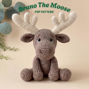 Op de afbeelding: Een gehaakt eland-speeltje in bruin met grote crèmekleurige geweien, donkere ogen en gestikte gezichtsdetails. De tekst "Bruno The Moose PDF PATTERN" staat in het groen boven het speeltje, dat in een zittende positie is afgebeeld.