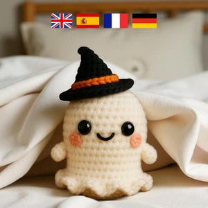 Könnte beinhalten: Eine gehäkelte Geisterpuppe mit einem schwarzen und orangefarbenen Hexenhut. Der Geist hat ein lächelndes Gesicht mit schwarzen Augen und rosigen Wangen. Die Puppe ist cremefarben und liegt auf einer weißen Decke.