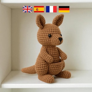May include: A handmade brown crochet kangaroo toy with a pouch, ears, and a tail. The stuffed animal is sitting on a white shelf. The kangaroo is a cute, soft toy.