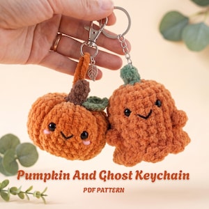 May include: Two crochet keychains featuring a round orange pumpkin with a brown stem and green leaf, and an orange ghost with a green top. Both have black eyes, stitched smiles, and pink cheeks. They are attached to silver metal key rings with a leaf charm.
