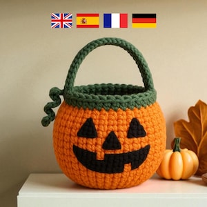 Puede incluir: Una cesta de calabaza de ganchillo naranja con un asa y borde verdes, y una cara de linterna tallada. La cesta está decorada con rasgos faciales negros. Una pequeña calabaza y una vela están en el fondo.