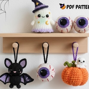 Op de afbeelding: Een verzameling handgemaakte gehaakte Halloween-decoraties. Inclusief een spook, twee oogbalornamenten, een vleermuis, een pompoen met een spook en een enkel oogbalornament. De decoraties worden tentoongesteld op een houten plank en haken.