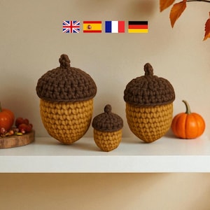 Könnte beinhalten: Drei gehäkelte Eichel-Dekorationen in verschiedenen Größen. Die Eichelkappen sind braun, die Körper gelb-orange. Ein kleiner Kürbis und eine brennende Kerze sind ebenfalls zu sehen.