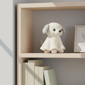 Könnte beinhalten: Eine weiße Häkelhundfigur mit grauen Ohren und einem kleinen Schwanz sitzt auf einem Holzregal. Ein gerahmtes Bild mit einem abstrakten Design befindet sich neben dem Hund. Das Regal ist Teil eines hellen Holzschranks.