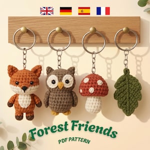 Könnte beinhalten: Vier gehäkelte Schlüsselanhänger an einer Holzleiste, bestehend aus einem Fuchs, einer Eule, einem rot-weißen Pilz und einem grünen Blatt. Der Text Forest Friends PDF PATTERN steht in Grün unten, darüber vier kleine Nationalflaggen.