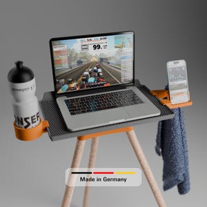 Könnte beinhalten: Ein grauer Laptop auf einem Stativständer mit einer orangefarbenen und grauen Ablage. Eine Wasserflasche und ein Smartphone sind am Ständer befestigt. Der Laptop-Bildschirm zeigt eine Radfahrsimulation. Ein blaues Handtuch hängt am Ständer.