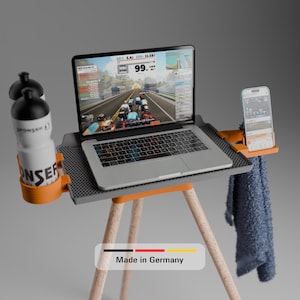 Könnte beinhalten: Ein grauer Laptopständer mit drei hellen Holzbeinen, ausgestattet mit einem orangefarbenen Flaschenhalter mit zwei weißen Wasserflaschen, einer orangefarbenen Smartphone-Halterung mit Handy und einer strukturierten Oberfläche. Ein blaues Handtuch hängt an der Seite. Der Text lautet Made in Germany.