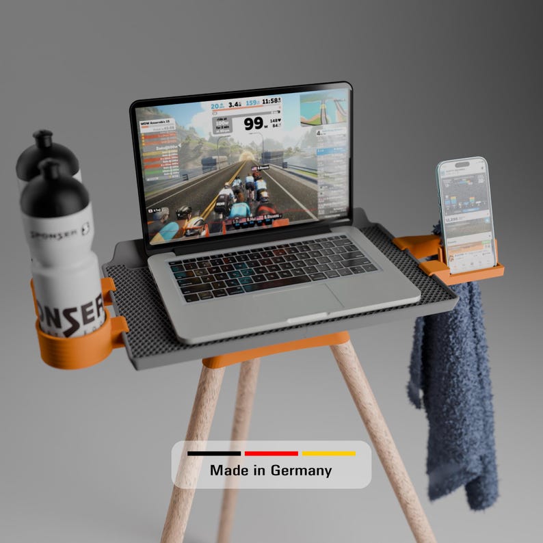 Peut inclure: Un support pour ordinateur portable gris avec trois pieds en bois clair, dot&eacute; d'un porte-bouteille orange avec deux bouteilles d'eau blanches, d'un support de smartphone orange avec un t&eacute;l&eacute;phone et d'une surface textur&eacute;e. Une serviette bleue est suspendue sur le c&ocirc;t&eacute;. Le texte indique Made in Germany.