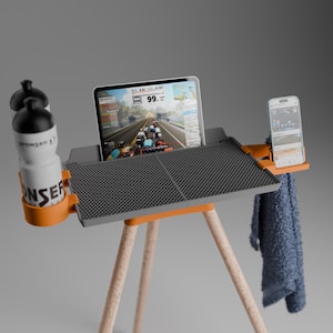 Peut inclure: Un bureau de cyclisme en salle gris avec une surface textur&eacute;e, des accents orange et des pieds en bois clair. Il comprend un support pour tablette affichant une application de cyclisme, deux bidons blancs, un support pour smartphone et une serviette bleue.