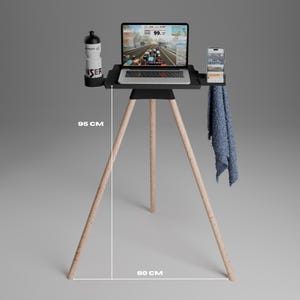 Peut inclure: Un support tr&eacute;pied noir avec des pieds en bois clair, mesurant 95 cm de hauteur et 80 cm de largeur. Le plateau sup&eacute;rieur accueille un ordinateur portable affichant une simulation cycliste, une gourde blanche, un smartphone et une serviette bleue.