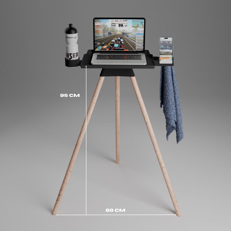 Peut inclure: Un support tr&eacute;pied noir avec des pieds en bois clair, mesurant 95 cm de hauteur et 80 cm de largeur. Le plateau sup&eacute;rieur accueille un ordinateur portable affichant une simulation cycliste, une gourde blanche, un smartphone et une serviette bleue.