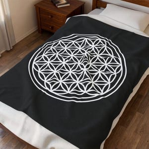May include: A black throw blanket featuring a large, centered white geometric pattern known as the flower of life. The design consists of multiple overlapping circles arranged in a hexagonal symmetry, creating a complex, repeating floral motif.