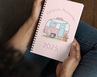 May include: A pink spiral-bound notebook with the words "Caravan Memories" and the year "2025" printed on the cover. The cover features a cartoon illustration of a pink and blue camper with a banner and potted flowers.
