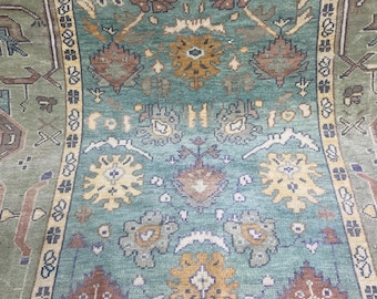 May include: A detailed, close-up view of a patterned rug. The rug features a central teal field with floral and geometric designs in shades of yellow, brown, and red. The border is a lighter green with repeating patterns.
