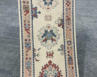 May include: A rectangular, cream-colored rug with an intricate pattern. The rug features a central design with floral and geometric motifs in shades of red, blue, and beige. The border is a mix of pink, blue, and red.