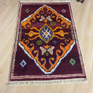 May include: A rectangular, hand-woven rug with a deep burgundy field and an intricate central design in orange, beige, and blue. The rug features a geometric pattern and a cream-colored fringe border. The rug is placed on a light wood floor.