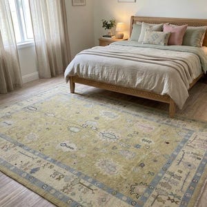 May include: A large, rectangular, distressed-look rug in a bedroom setting. The rug features a muted yellow-green field with a detailed border in blue and beige. The room includes a wooden bed frame and neutral-toned bedding.