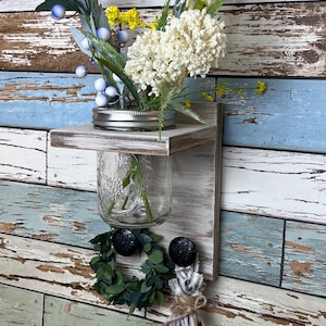 May include: A rustic wooden shelf with a mason jar vase holding a bouquet of white and yellow flowers. The shelf has two black knobs and a small green wreath hanging below. The shelf is mounted on a weathered wood background.