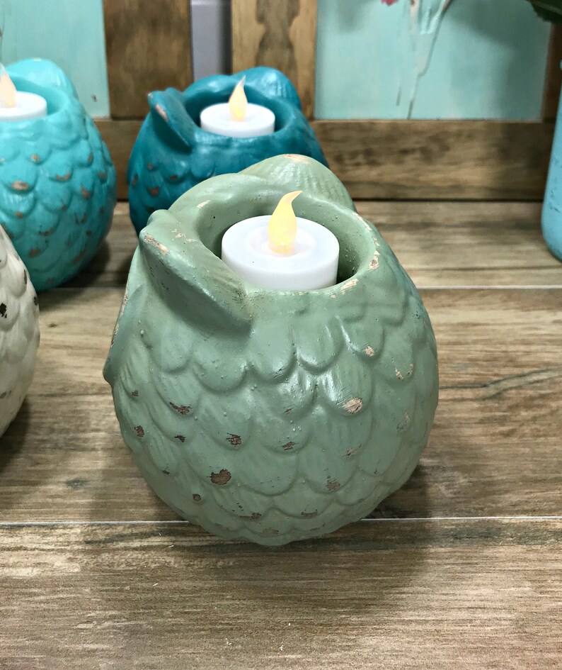 Puede incluir: Un conjunto de portavelas de cer&aacute;mica en forma de b&uacute;ho de color verde azulado y verde con un acabado desgastado. Cada b&uacute;ho tiene una peque&ntilde;a abertura en la parte superior para sostener una vela de t&eacute;.