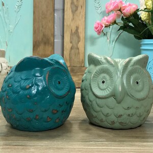 Puede incluir: Dos figuritas de b&uacute;ho de cer&aacute;mica, una azul verdosa y otra verde claro, con detalles texturizados y ojos pintados.