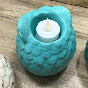 Puede incluir: Portavelas de cer&aacute;mica azul turquesa en forma de b&uacute;ho con una vela de t&eacute; blanca en su interior. El b&uacute;ho tiene una superficie texturizada que se asemeja a escamas.