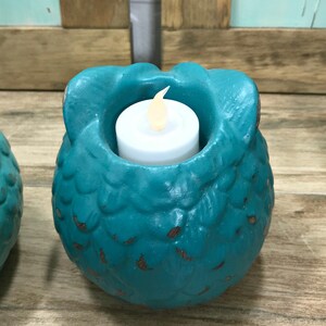 Puede incluir: Portavelas de cer&aacute;mica de color turquesa en forma de b&uacute;ho con una vela de t&eacute; blanca en su interior. El b&uacute;ho tiene una superficie texturizada y est&aacute; sentado sobre una superficie de madera.