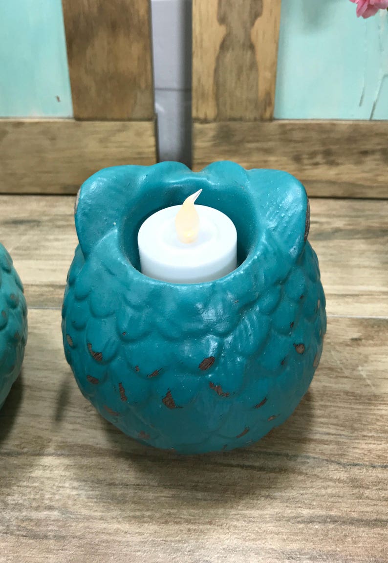 Puede incluir: Portavelas de cer&aacute;mica de color turquesa en forma de b&uacute;ho con una vela de t&eacute; blanca en su interior. El b&uacute;ho tiene una superficie texturizada y est&aacute; sentado sobre una superficie de madera.