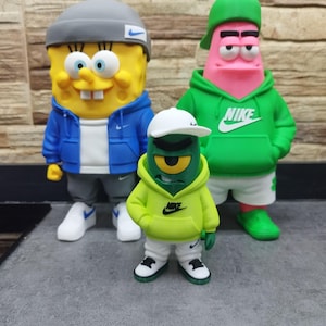 May include: Three cartoon character figurines wearing Nike apparel. A yellow sponge figure in a gray beanie and blue jacket, a pink starfish figure in a green hoodie and cap, and a small green cyclops figure in a lime hoodie and white cap.