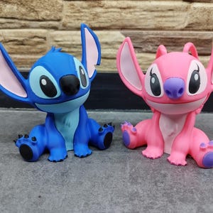 May include: Two figurines of the characters Stitch and Angel from the animated film Lilo & Stitch. The blue Stitch figure has large ears with pink insides, and the pink Angel figure has similar features. Both have large eyes and are in a sitting position.