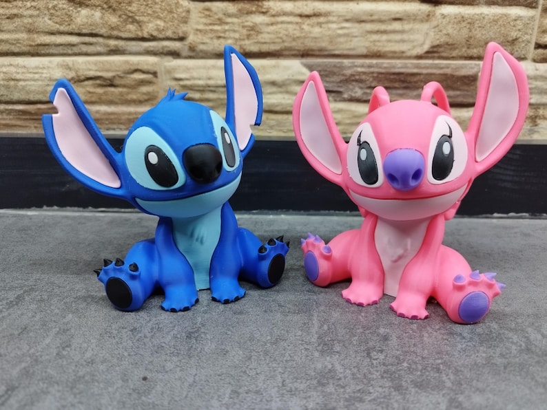 May include: Two figurines of the characters Stitch and Angel from the animated film Lilo & Stitch. The blue Stitch figure has large ears with pink insides, and the pink Angel figure has similar features. Both have large eyes and are in a sitting position.