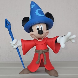 May include: A figurine of Mickey Mouse dressed as a sorcerer, wearing a blue hat with a crescent moon and stars, and a red robe. He holds a blue staff and has white gloves. The figure is standing with one arm raised.
