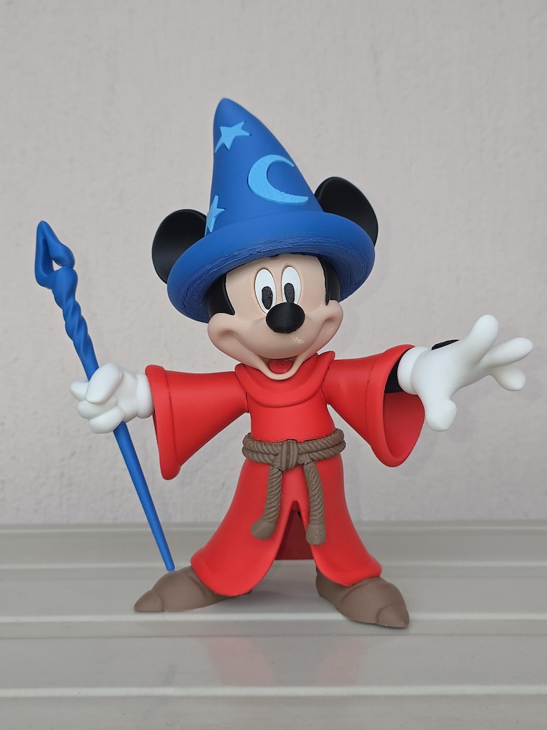 May include: A figurine of Mickey Mouse dressed as a sorcerer, wearing a blue hat with a crescent moon and stars, and a red robe. He holds a blue staff and has white gloves. The figure is standing with one arm raised.