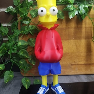 May include: A yellow cartoon character figurine with spiky hair and large white eyes. The figure wears a bright red hooded sweatshirt, blue shorts, and blue sneakers with white laces and soles. The character stands upright with hands in pockets.
