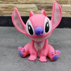 May include: A pink figurine of the character Angel from the Lilo & Stitch franchise. The figurine has large ears, a purple nose, and purple accents on its feet. The figure is sitting down with its arms and legs outstretched.