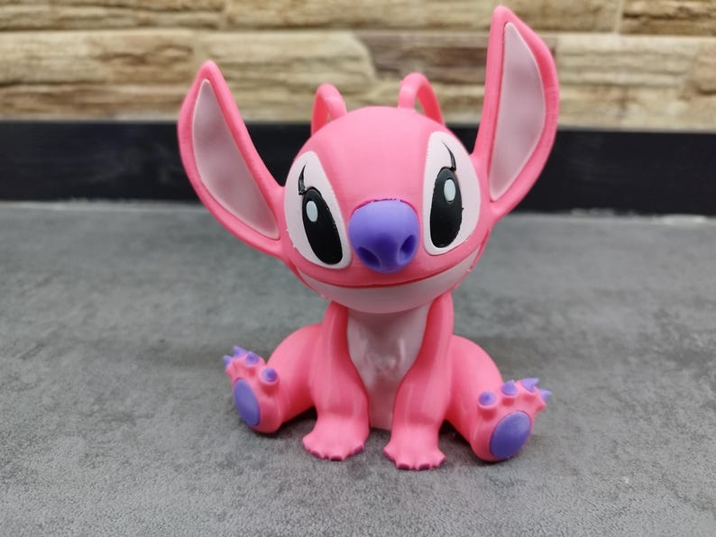 May include: A pink figurine of the character Angel from the Lilo & Stitch franchise. The figurine has large ears, a purple nose, and purple accents on its feet. The figure is sitting down with its arms and legs outstretched.