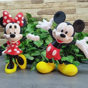 May include: Two figurines of Mickey and Minnie Mouse. Minnie wears a red dress with white polka dots and a matching bow. Mickey is in red shorts with white buttons. Both have yellow shoes and white gloves.