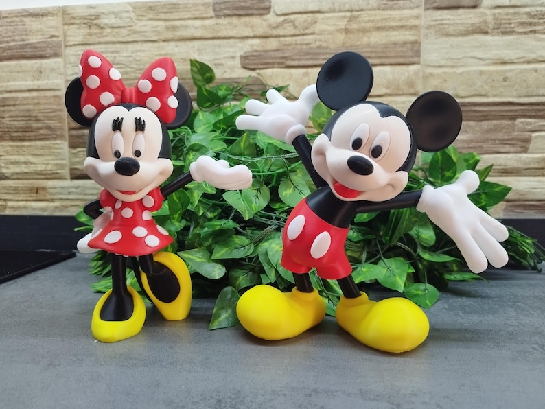 May include: Two figurines of Mickey and Minnie Mouse. Minnie wears a red dress with white polka dots and a matching bow. Mickey is in red shorts with white buttons. Both have yellow shoes and white gloves.