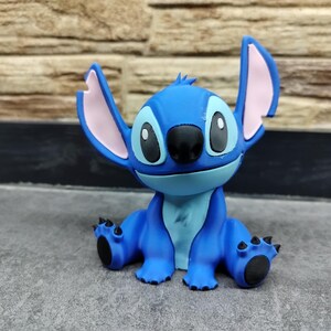 May include: A blue figurine of the Disney character Stitch, with large ears, black eyes, and a black nose. The figurine is sitting on a gray surface, with a brick wall in the background. The figurine is a popular character from the Lilo & Stitch franchise.