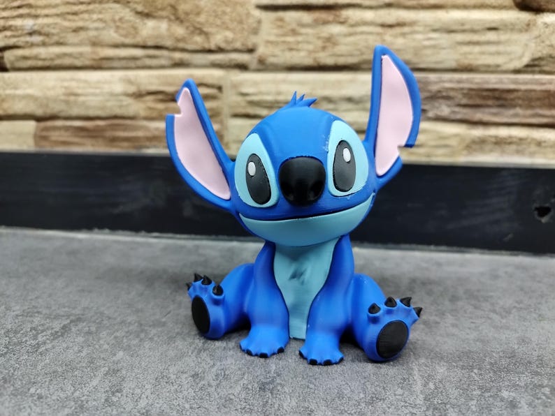 May include: A blue figurine of the Disney character Stitch, with large ears, black eyes, and a black nose. The figurine is sitting on a gray surface, with a brick wall in the background. The figurine is a popular character from the Lilo & Stitch franchise.