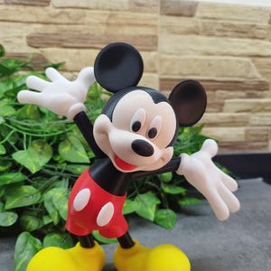 May include: A figurine of Mickey Mouse, a classic cartoon character, is depicted with outstretched white gloved hands. Mickey wears red shorts with white buttons, yellow shoes, and a black body and ears. The figurine is set against a backdrop of green leaves and a textured wall.